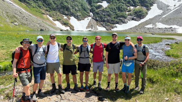 Neun Männer in Wanderkleidung stehen Arm in Arm vor einem Bergsee mit Schneefeldern und grünen Hängen